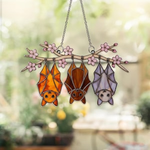 K&ouml;nnte beinhalten: Buntglas-Suncatcher mit drei Flederm&auml;usen, die an einem Ast mit rosa Bl&uuml;ten h&auml;ngen. Die Flederm&auml;use sind in Orange-, Braun- und Lavendelt&ouml;nen gehalten. Der Suncatcher h&auml;ngt an einer silbernen Kette, ideal f&uuml;r Fenster- oder Wanddekoration.