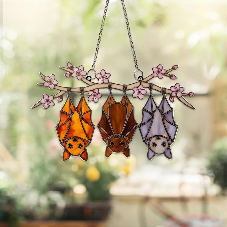 K&ouml;nnte beinhalten: Buntglas-Suncatcher mit drei Flederm&auml;usen, die an einem Ast mit rosa Bl&uuml;ten h&auml;ngen. Die Flederm&auml;use sind in Orange-, Braun- und Lavendelt&ouml;nen gehalten. Der Suncatcher h&auml;ngt an einer silbernen Kette, ideal f&uuml;r Fenster- oder Wanddekoration.