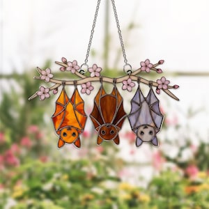 K&ouml;nnte beinhalten: Buntglas-Fensterdekoration mit drei Flederm&auml;usen in Orange, Braun und Lavendel, die an einem Ast mit rosa Bl&uuml;ten h&auml;ngen. Die Flederm&auml;use h&auml;ngen kopf&uuml;ber, mit einer Kette. Ein dekoratives St&uuml;ck f&uuml;r Halloween oder das ganze Jahr.