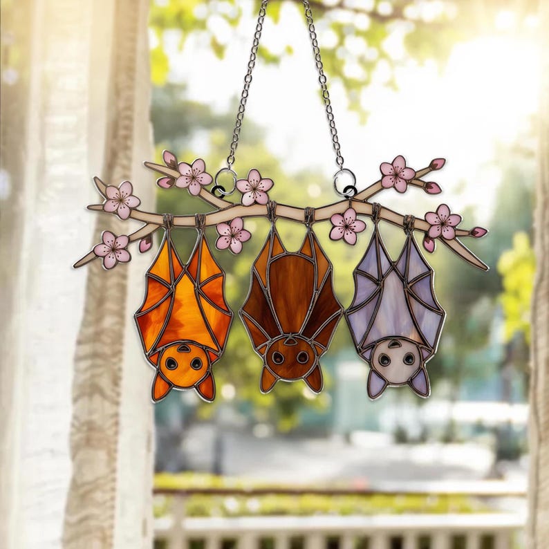 K&ouml;nnte beinhalten: Buntglas-Fensterdekoration mit drei Flederm&auml;usen in Orange, Braun und Lavendel, die an einem Ast mit rosa Bl&uuml;ten h&auml;ngen. Die Flederm&auml;use sind an einer silbernen Kette aufgeh&auml;ngt.