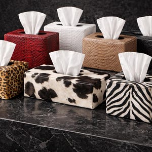 May include: A selection of tissue box covers with animal print and textured finishes. Designs include leopard print, zebra stripes, and cowhide, alongside faux leather in red, brown, white, and black. Each box contains a fan of white tissues.