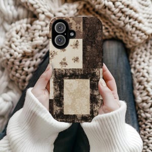 Könnte beinhalten: Eine rechteckige Smartphone-Hülle mit einem Patchwork-Design aus dunkelbraunen und cremefarbenen Feldern. Das Muster wechselt zwischen einfarbigen dunkelbraunen Texturen und cremefarbenen Abschnitten, die mit kleinen, braunen Blumenmotiven verziert sind.