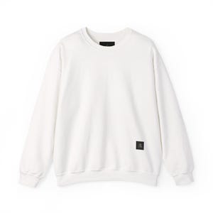 Pode incluir: Um moletom branco com gola redonda e mangas compridas. O moletom tem um pequeno patch retangular preto no lado inferior esquerdo e uma pequena etiqueta preta na parte de trás do pescoço. O moletom é feito de material macio.