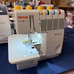 May include: A white and gray Feiyue FY525 serger sewing machine with yellow, green, blue, and red thread guides. The machine has a white work surface and the text "FEIYUE FABRIC MOUSE" is visible on the front.