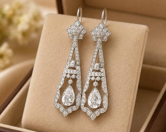 May include: A pair of silver-toned drop earrings featuring an intricate geometric frame encrusted with small clear stones. Each earring terminates in a single pear-shaped clear stone suspended within the center of the openwork metal design.