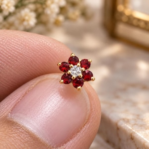 May include: A small gold stud earring shaped like a flower, featuring six deep red circular gemstones arranged as petals around a single clear, brilliant-cut white gemstone at the center, all set in a polished gold metal frame.