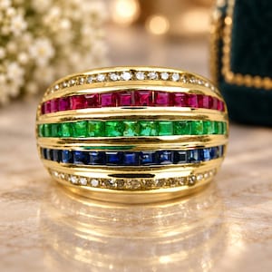 May include: A gold band ring featuring four horizontal rows of channel-set gemstones. The top and bottom rows consist of small clear diamonds, while the middle rows contain square-cut vibrant red rubies, bright green emeralds, and deep blue sapphires.