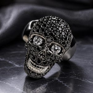 May include: A skull-shaped ring featuring a polished silver metal band and a skull head encrusted with numerous small black stones. The eye sockets of the skull are set with two large, clear, round-cut stones, and the teeth are made of silver.