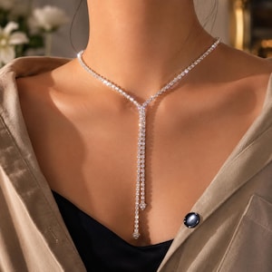 May include: A silver-toned lariat necklace featuring a continuous strand of clear, round-cut stones. The design includes a circular collar section that transitions into two parallel, dangling vertical chains, each finished with a small, pointed stone accent.