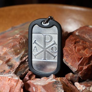 May include: A rectangular silver-toned metal dog tag pendant featuring an engraved Chi Rho symbol with the Greek letters Alpha and Omega on either side. The pendant is encased in a black rubber silencer border and includes a small metal ball chain.