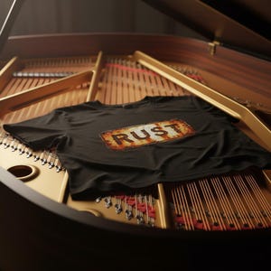 May include: Black t-shirt with the word "RUST" in a distressed, rusty-looking design. The shirt is lying on the strings of a grand piano. The piano's wood and gold accents provide a warm contrast to the shirt's dark color.