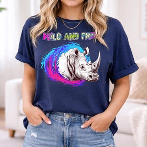 Peut inclure: T-shirt bleu marine avec un motif de rhinocéros et le texte "WILD AND FREE". Le rhinocéros est entouré d'un tourbillon de bleu, de rose et de violet. Le t-shirt a des manches retroussées.