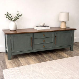 May include: A green and brown wooden sideboard with three drawers and two doors. The doors have vertical paneling and bronze handles. A white vase with greenery, books, a bowl, and a lamp sit on top. The sideboard is on a wood floor.