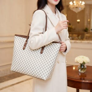 May include: A cream tote bag featuring a geometric pattern in grey and brown. It has brown handles and trim, with gold rivets. The bag is held by a person wearing a cream coat.