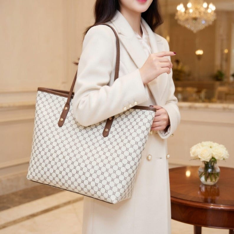 May include: A cream tote bag featuring a geometric pattern in grey and brown. It has brown handles and trim, with gold rivets. The bag is held by a person wearing a cream coat.