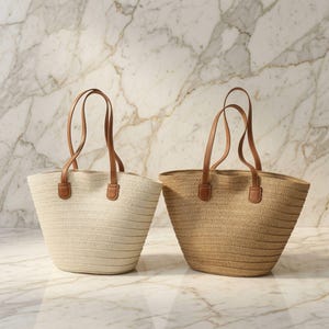 May include: Two tote bags with a woven straw-like texture. One bag is off-white and the other is a natural tan color. Both bags feature two thin, brown leather-like shoulder straps attached with rectangular tabs at the top edge of the bag.