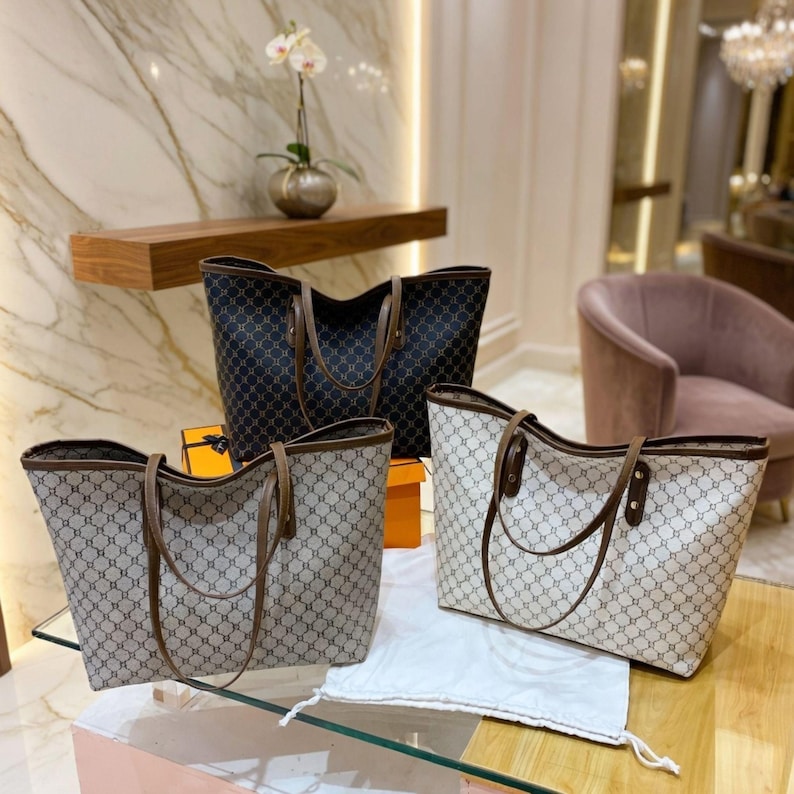 May include: Four tote bags are displayed. Three bags feature a light beige pattern with brown handles and trim. The fourth bag is black with a similar pattern and brown handles. The bags are on a pink surface.