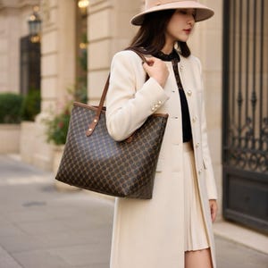 May include: A brown tote bag with a repeating geometric pattern. The bag has brown leather handles and trim. The person is wearing a cream-coloured coat and a beige hat.