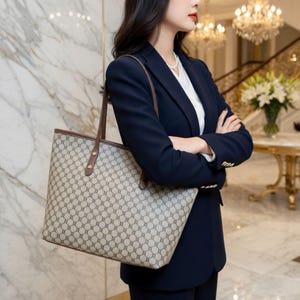 May include: A beige tote bag with a repeating geometric pattern and brown leather handles. The bag is held by a person wearing a navy blue blazer and white shirt. The bag has a rectangular shape.