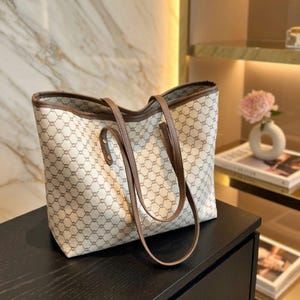 May include: Beige tote bag with a repeating geometric pattern and brown leather handles and trim. The bag is sitting on a dark surface.