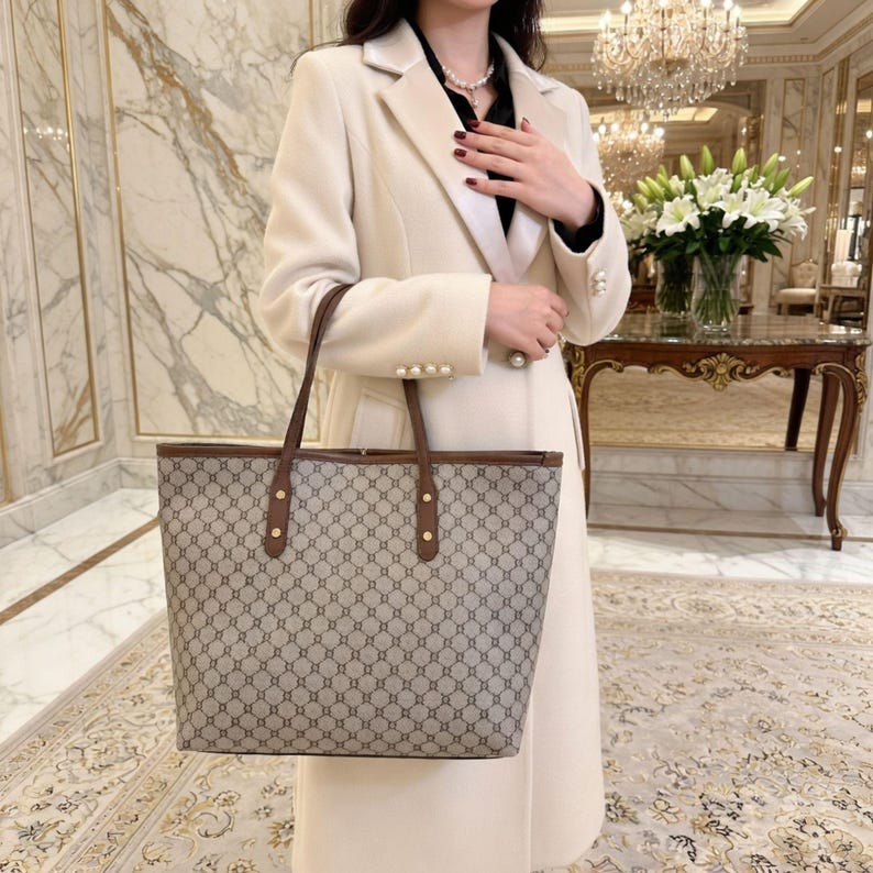 May include: A beige tote bag with a brown geometric pattern and brown leather handles. The bag has a rectangular shape and is held by a person wearing a cream-coloured coat.