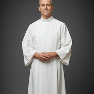 May include: A white clergy robe with a high collar and wide sleeves. The robe is long and flowing, with vertical seams down the front. The person is standing in front of a gray background.