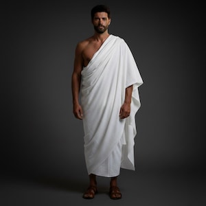 Op de afbeelding: Een persoon in een witte toga, gedrapeerd over de linkerschouder en om het lichaam. De persoon draagt bruine sandalen. De achtergrond is effen grijs. De toga is een traditioneel kledingstuk.