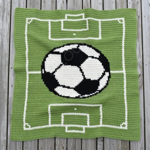 May include: A handmade crochet blanket featuring a soccer field design. The blanket has a green background with white lines depicting the field markings and a large soccer ball in the center. The ball is black and white.