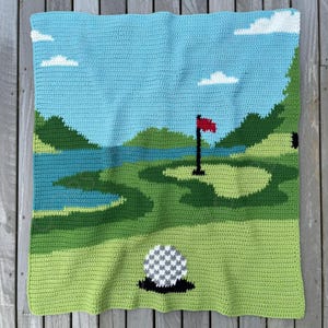 Könnte beinhalten: Eine gehäkelte Decke mit einer Golfplatzszene. Das Design zeigt einen blauen Himmel mit weißen Wolken, grüne Fairways, einen See und einen Golfball. Eine rote Fahne markiert das Loch. Die Decke ist in verschiedenen Grün- und Blautönen gehalten.