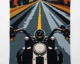 May include: A rectangular crochet tapestry featuring a first-person perspective of a motorcycle riding down a road. The scene includes a black motorcycle dashboard, handlebars, and a headlight, with a yellow centre line and autumn trees.