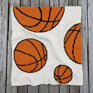 May include: A crocheted blanket featuring a repeating pattern of orange basketballs with black lines on a white background. The blanket is square and appears to be handmade, with a textured stitch.
