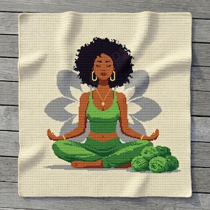 May include: A square crochet blanket featuring a pixelated design of a person with dark skin and curly hair sitting in a lotus pose. The person wears a green top and trousers, gold hoop earrings, and a necklace. A grey flower shape is behind them.