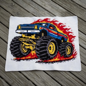 May include: A white crocheted square featuring a colorful monster truck design. The truck is blue with yellow and red accents, large black tires, and flames. The design is centered on the white background.