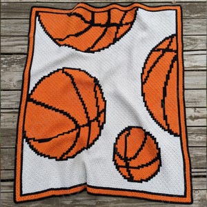 Puede incluir: Una manta cuadrada de ganchillo hecha a mano con un diseño de baloncesto. La manta tiene un fondo blanco con balones de baloncesto naranjas delineados en negro. El borde es naranja con un ribete negro.