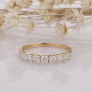 May include: A gold band ring featuring a row of seven round, clear, faceted stones set in a shared prong setting. The metal has a polished finish, and the stones are arranged horizontally across the front arc of the thin, circular band.