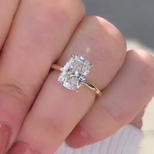 May include: A gold ring featuring a large, rectangular cushion-cut clear gemstone set in a four-prong setting. The band is thin, polished, and metallic gold, showcasing a classic solitaire design with a faceted stone that reflects light.
