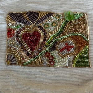May include: A rectangular beaded artwork featuring a central red heart, surrounded by gold, brown, and green beads. The design includes floral and abstract elements, with a textured appearance. The artwork is set against a neutral fabric background.