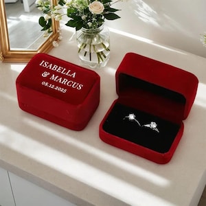 Op de afbeelding: Twee diamanten verlovingsringen in een rode fluwelen ringdoos. De open doos onthult de ringen tegen een zwart interieur. Een gesloten rode fluwelen doos met de namen "ISABELLA & MARCUS" en de datum "08.12.2025" is ook aanwezig.