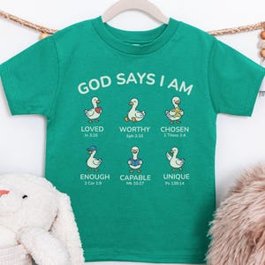 May include: A teal-colored t-shirt with the text "GOD SAYS I AM" in white. Below the text are six cartoon ducks with various accessories and the words "LOVED," "WORTHY," "CHOSEN," "ENOUGH," "CAPABLE," and "UNIQUE."