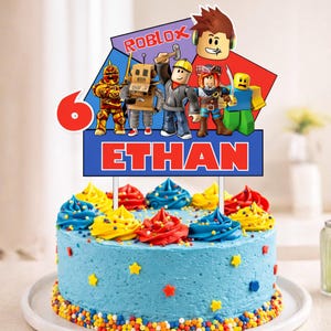 K&ouml;nnte beinhalten: Ein blau glasierter Kuchen, verziert mit bunten Zuckergusswirbeln und Streuseln. Ein Tortenaufsatz zeigt Roblox-Charaktere, die Zahl 6 und den Namen "ETHAN" in roten Buchstaben. Der Kuchen steht auf einem wei&szlig;en Teller.