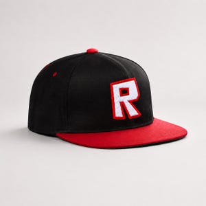 Puede incluir: Una gorra de béisbol negra con una letra R blanca destacada con borde rojo bordada en el panel frontal. La gorra tiene una visera plana roja y un pequeño botón rojo en la parte superior central, con un diseño estructurado de seis paneles.
