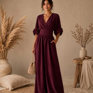 May include: A deep burgundy jumpsuit featuring a V-neckline, a wide gathered waistband, and three-quarter length sleeves with gathered cuffs. The garment has a floor-length wide-leg silhouette and includes side pockets, paired with a small woven bag.