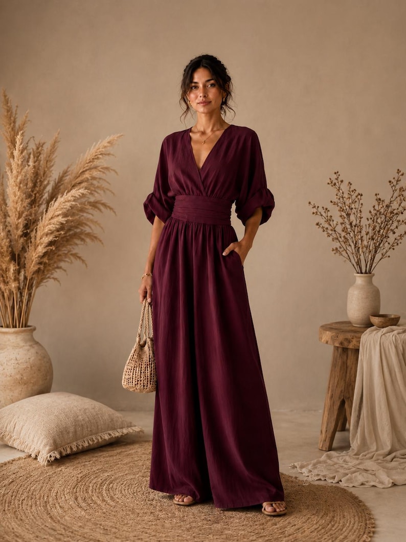 May include: A deep burgundy jumpsuit featuring a V-neckline, a wide gathered waistband, and three-quarter length sleeves with gathered cuffs. The garment has a floor-length wide-leg silhouette and includes side pockets, paired with a small woven bag.