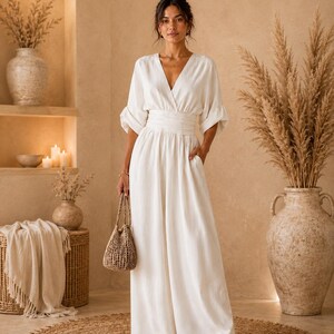 May include: A floor-length off-white jumpsuit featuring a deep V-neckline, elbow-length sleeves with gathered cuffs, and a wide, ruched waistband. The garment includes side pockets and wide-leg trousers, paired with a small, woven tan handbag.