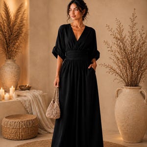 May include: A black maxi dress featuring a V-neckline, elbow-length sleeves with cuffed hems, and a wide, gathered waistband. The dress has a long, flowing skirt with side pockets and is paired with a small, woven, light-coloured handbag.