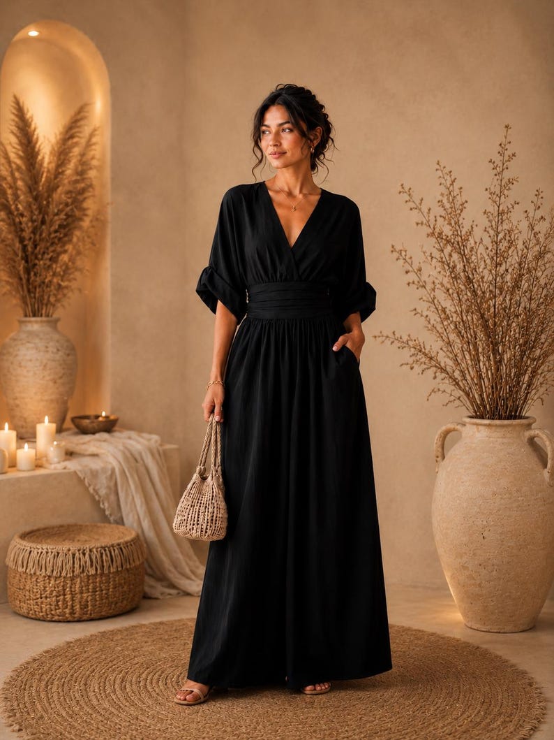 May include: A black maxi dress featuring a V-neckline, elbow-length sleeves with cuffed hems, and a wide, gathered waistband. The dress has a long, flowing skirt with side pockets and is paired with a small, woven, light-coloured handbag.