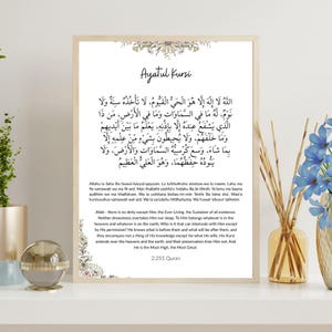 Puede incluir: Una lámina de arte mural enmarcada que presenta el Ayatul Kursi en caligrafía árabe, seguida de su transliteración y traducción al inglés. El diseño incluye delicadas ilustraciones florales en la parte superior e inferior sobre un fondo blanco.
