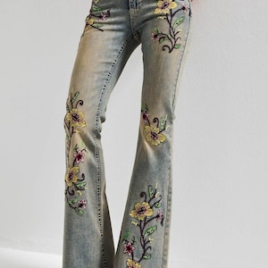 Könnte beinhalten: Hellblaue Schlagjeans aus Denim mit gestickten Blumenmustern in Gelb, Pink und Grün. Die Jeans verfügt über ein klassisches Fünf-Taschen-Design mit Knopf- und Reißverschluss sowie detaillierte Nähte entlang der Beine.