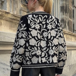May include: Black jacket with intricate floral embroidery in a cream color. The jacket features a crew neck and long sleeves. The floral design covers the entire back and sleeves of the jacket. The jacket is paired with black shorts.