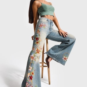 May include: Light blue denim jeans featuring a high-waisted fit and wide-leg silhouette. The jeans are decorated with vibrant, multi-colored floral embroidery running down the side of the right leg and a smaller floral motif on the left leg.